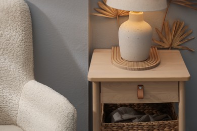 Photo of Stylish wooden side table with lamp near armchair at home, closeup