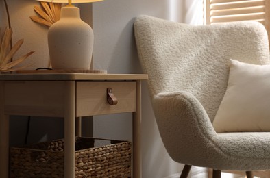 Photo of Stylish wooden side table with lamp near armchair at home, closeup