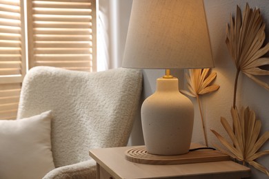 Photo of Stylish wooden side table with lamp near armchair at home, closeup