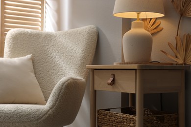 Photo of Stylish wooden side table with lamp near armchair at home, closeup
