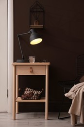 Photo of Wooden side table with glass of water, lamp and glasses near brown wall indoors