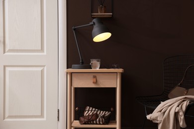 Photo of Wooden side table with glass of water, lamp and glasses near brown wall indoors