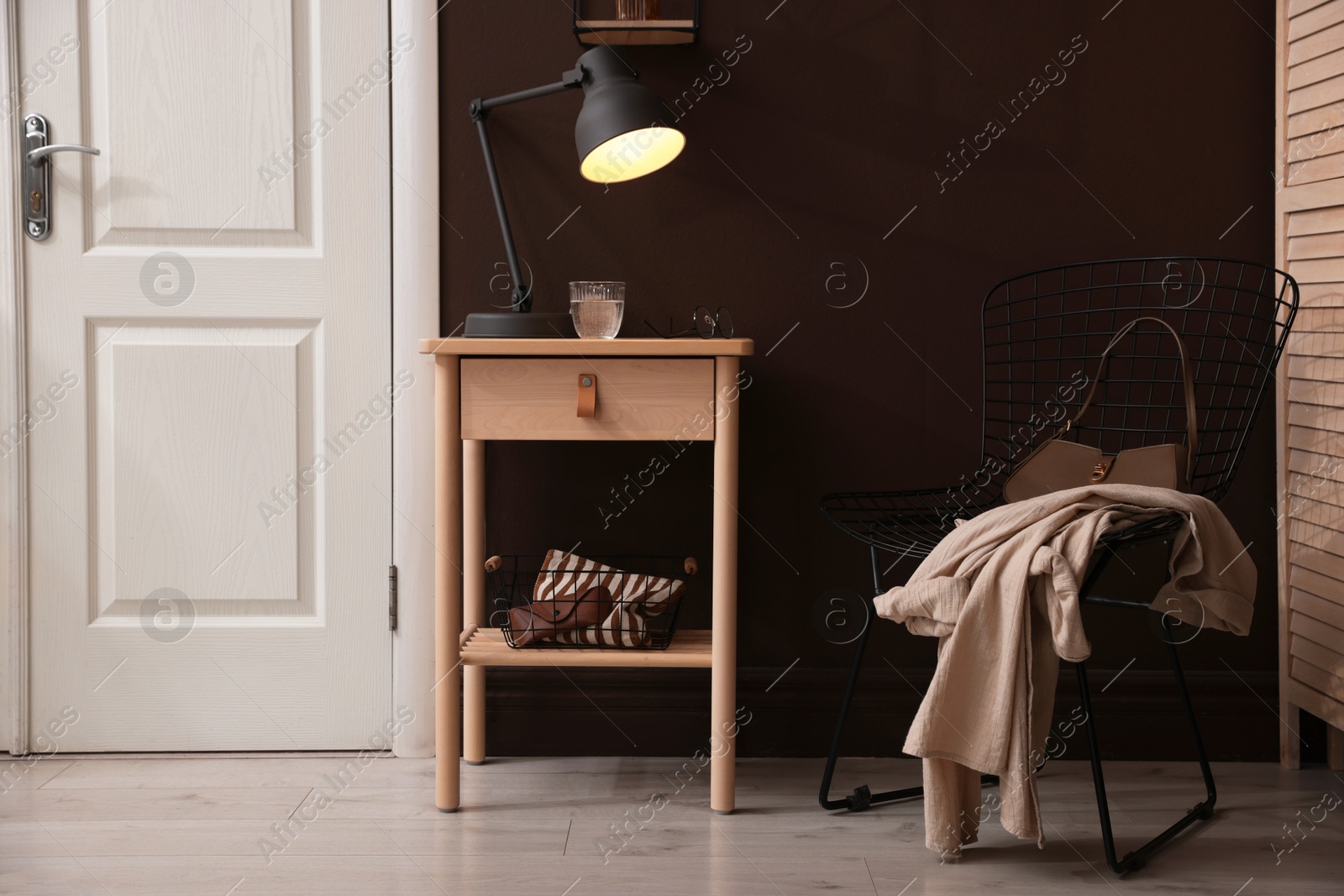 Photo of Wooden side table with glass of water, lamp and glasses near brown wall indoors