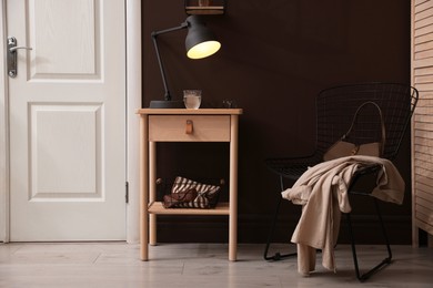 Photo of Wooden side table with glass of water, lamp and glasses near brown wall indoors