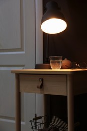 Photo of Wooden side table with glass of water, lamp and glasses near brown wall indoors