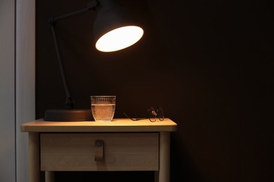 Photo of Wooden side table with glass of water, lamp and glasses near brown wall indoors, closeup. Space for text