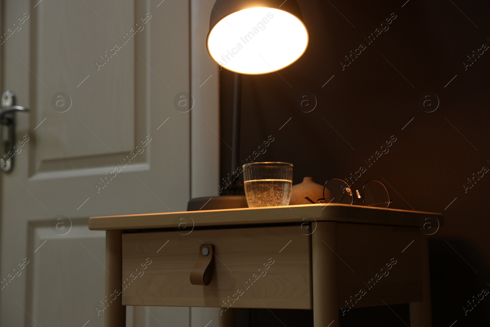 Photo of Wooden side table with glass of water, lamp and glasses near brown wall indoors, closeup