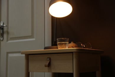 Photo of Wooden side table with glass of water, lamp and glasses near brown wall indoors, closeup