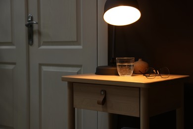 Photo of Wooden side table with glass of water, lamp and glasses near brown wall indoors, closeup. Space for text
