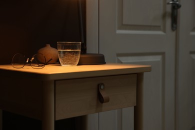 Photo of Wooden side table with glass of water and glasses near brown wall indoors, closeup