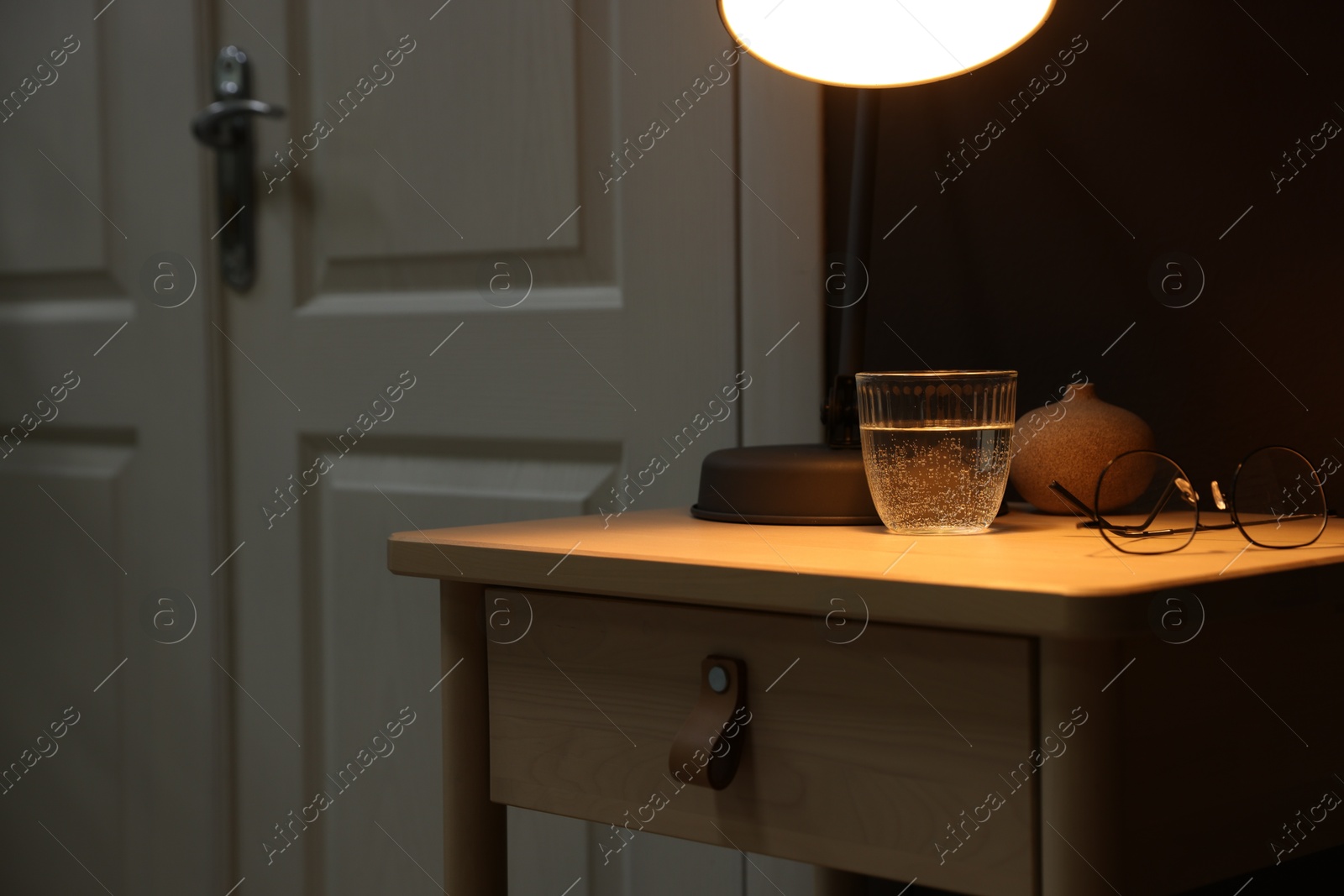 Wooden side table with glass of water, lamp and glasses near brown wall indoors, closeup. Space for text Photo of Wooden side table with glass of water, lamp and glasses near brown wall indoors, closeup. Space for text