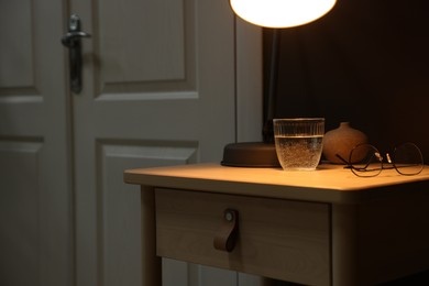 Photo of Wooden side table with glass of water, lamp and glasses near brown wall indoors, closeup. Space for text