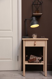 Photo of Wooden side table with glass of water, lamp and glasses near brown wall indoors