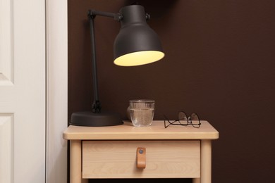 Photo of Wooden side table with glass of water, lamp and glasses near brown wall indoors