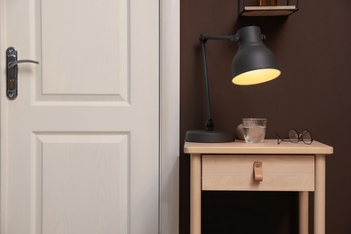 Photo of Wooden side table with glass of water, lamp and glasses near brown wall indoors