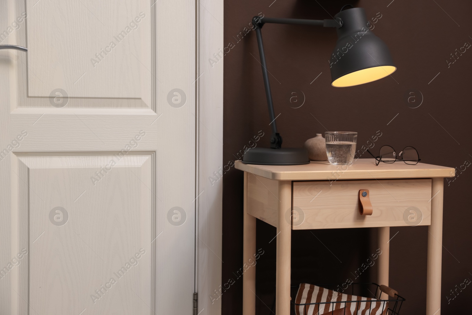 Photo of Wooden side table with glass of water, lamp and glasses near brown wall indoors