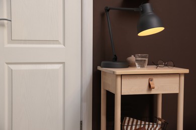 Photo of Wooden side table with glass of water, lamp and glasses near brown wall indoors