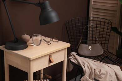 Photo of Wooden side table with glass of water, lamp and glasses near brown wall indoors