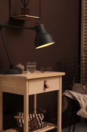 Photo of Wooden side table with glass of water, lamp and glasses near brown wall indoors