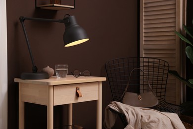 Photo of Wooden side table with glass of water, lamp and glasses near brown wall indoors