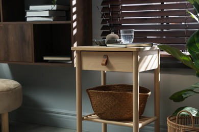 Photo of Wooden side table with glass of water, notebook, air reed diffuser and basket near window indoors