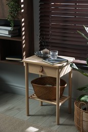 Photo of Wooden side table with glass of water, notebook, air reed diffuser and basket near window indoors