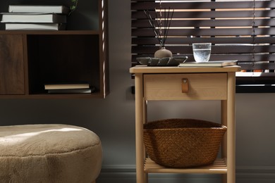 Photo of Wooden side table with glass of water, notebook, air reed diffuser and basket near window indoors