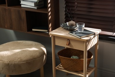 Photo of Wooden side table with glass of water, notebook, air reed diffuser and basket near window indoors