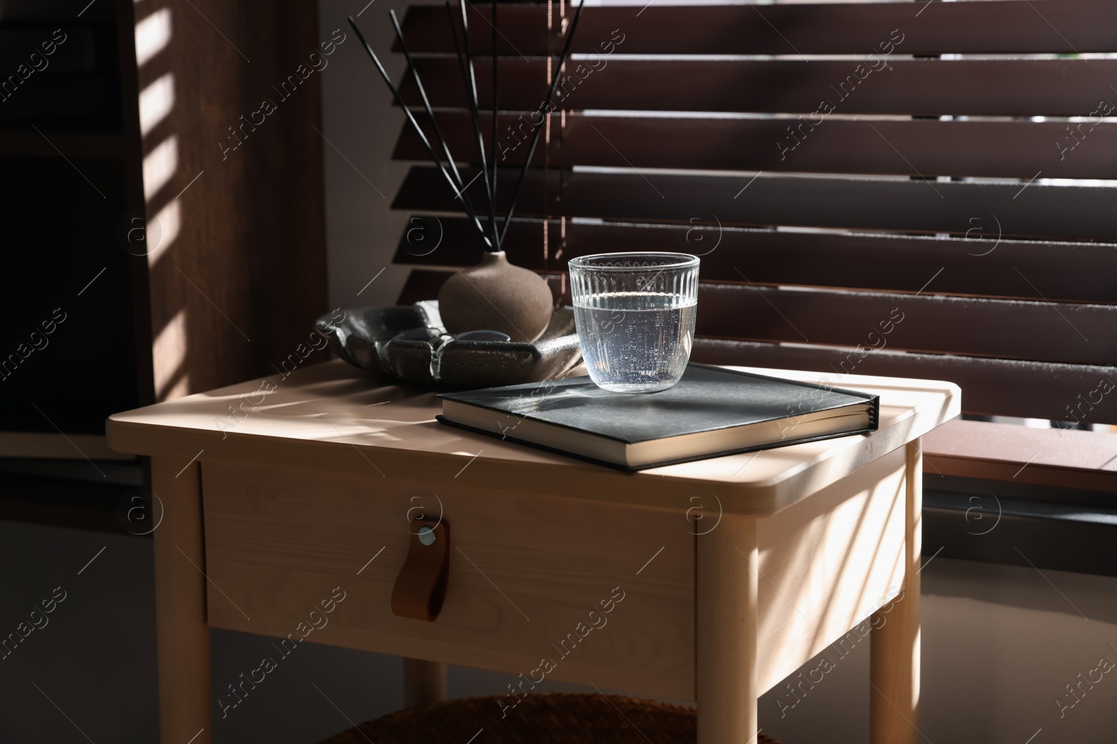 Wooden side table with glass of water, notebook and air reed diffuser indoors Photo of Wooden side table with glass of water, notebook and air reed diffuser indoors