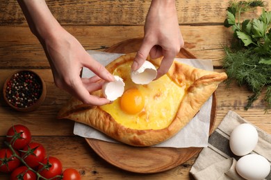 Photo of Woman breaking egg into delicious khachapuri at wooden table, top view