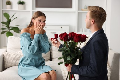 Photo of Man with engagement ring and bouquet of roses making marriage proposal to his excited girlfriend at home