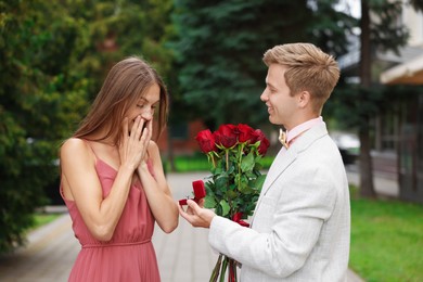 Photo of Smiling man with engagement ring and bouquet of roses making marriage proposal to his surprised girlfriend outdoors