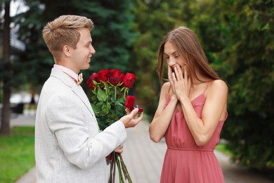 Photo of Smiling man with engagement ring and bouquet of roses making marriage proposal to his surprised girlfriend outdoors