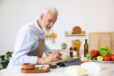Photo of Elderly man using tablet while cooking at white marble table in kitchen