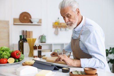 Photo of Elderly man using tablet while cooking at white marble table in kitchen