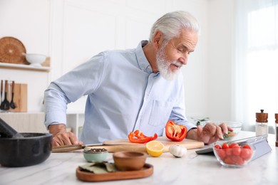 Photo of Elderly man using tablet while cooking at white marble table in kitchen