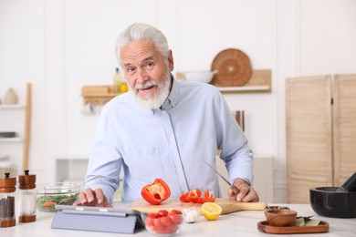 Photo of Elderly man using tablet while cooking at white marble table in kitchen