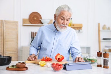 Photo of Elderly man using tablet while cooking at white marble table in kitchen