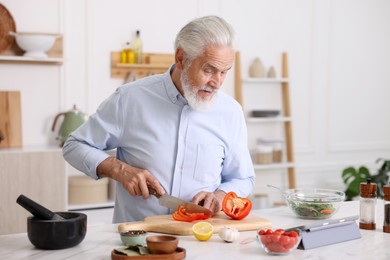 Photo of Elderly man using tablet while cooking at white marble table in kitchen