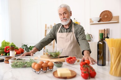 Photo of Elderly man cooking salad at white marble table in kitchen