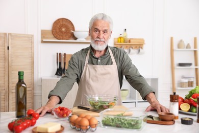 Photo of Elderly man cooking salad at white marble table in kitchen