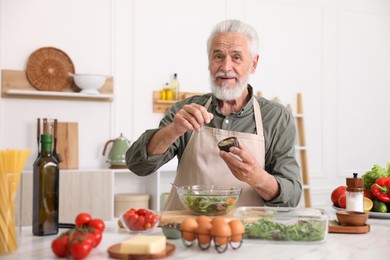 Photo of Elderly man cooking salad at white marble table in kitchen