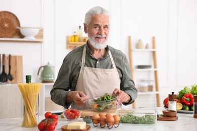 Photo of Elderly man cooking salad at white marble table in kitchen