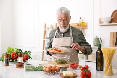 Photo of Elderly man cooking salad at white marble table in kitchen