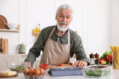 Photo of Elderly man using tablet while cooking at white marble table in kitchen