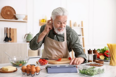 Photo of Elderly man using tablet while cooking at white marble table in kitchen