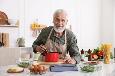 Photo of Elderly man using tablet while cooking at white marble table in kitchen