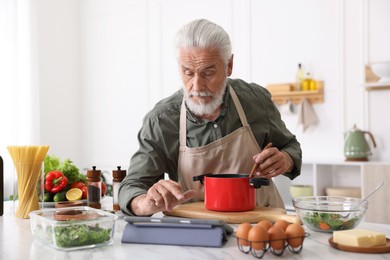 Photo of Elderly man using tablet while cooking at white marble table in kitchen