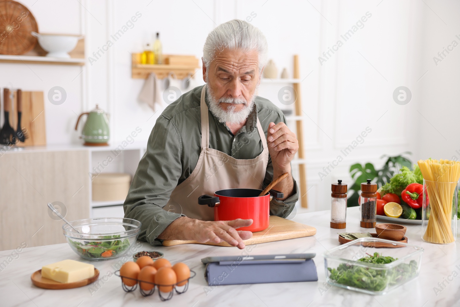 Elderly man using tablet while cooking at white marble table in kitchen Photo of Elderly man using tablet while cooking at white marble table in kitchen