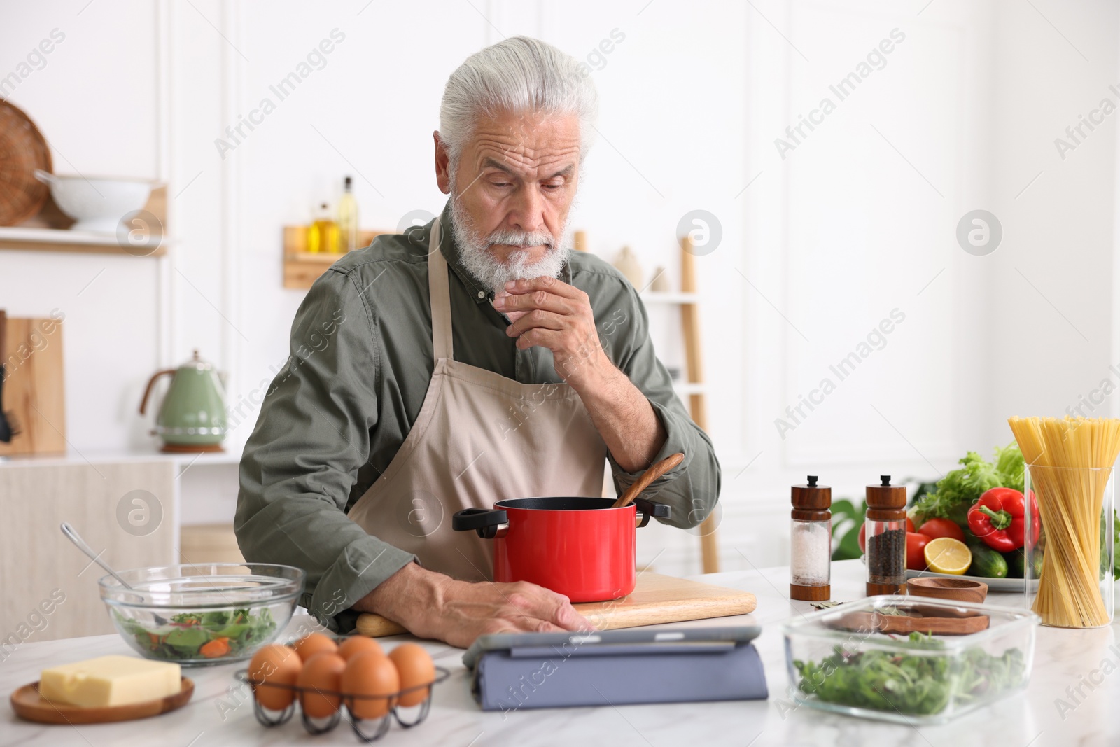 Elderly man using tablet while cooking at white marble table in kitchen Photo of Elderly man using tablet while cooking at white marble table in kitchen